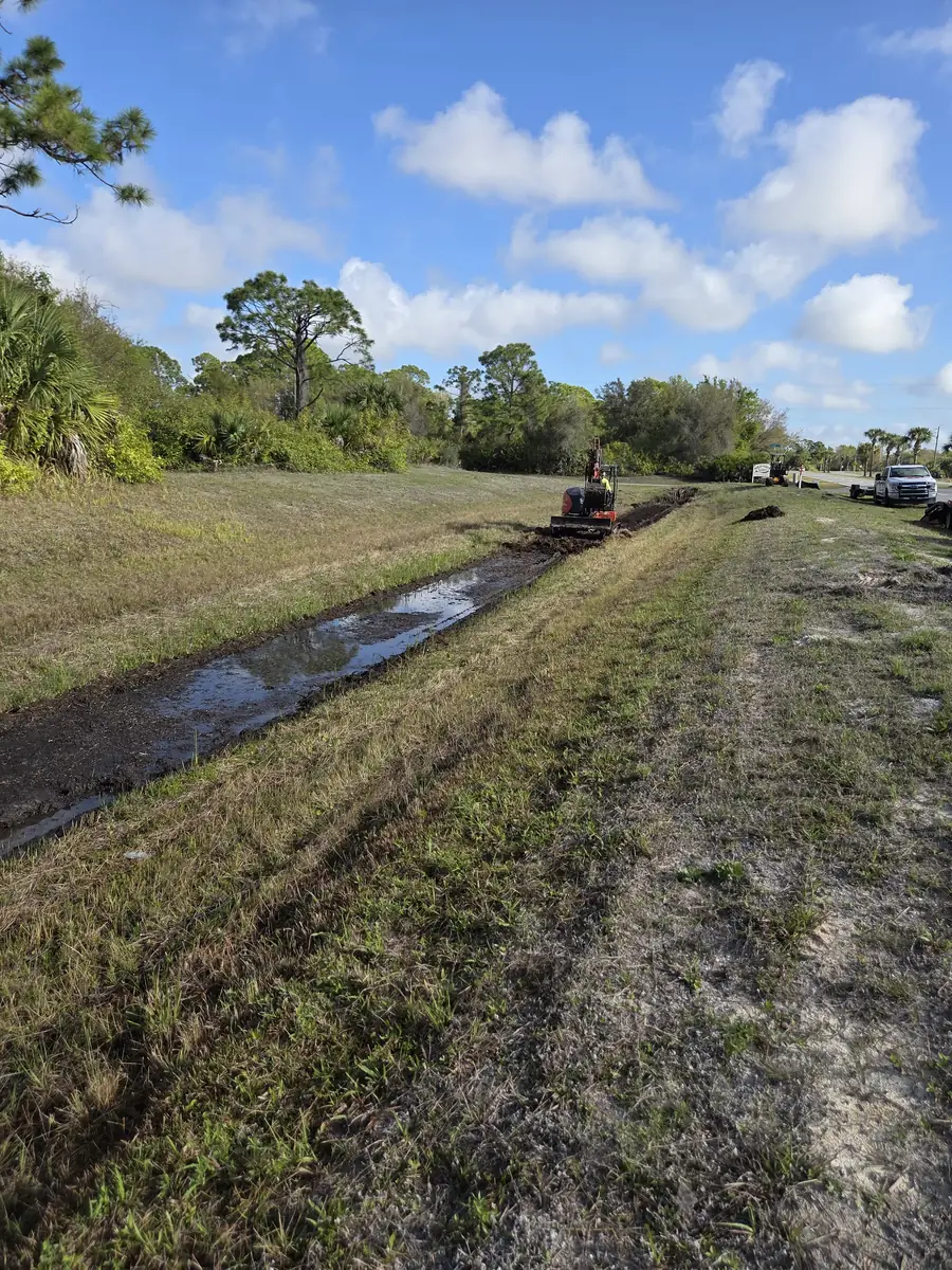 Long grassed swale re-cut back to grade with water standing in the new channel and the excavator working up ahead