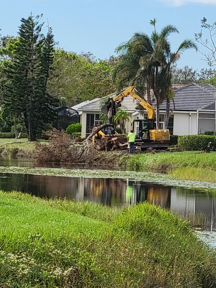 Excavator lifting a fallen tree and storm debris off a pond bank behind a home in a residential community