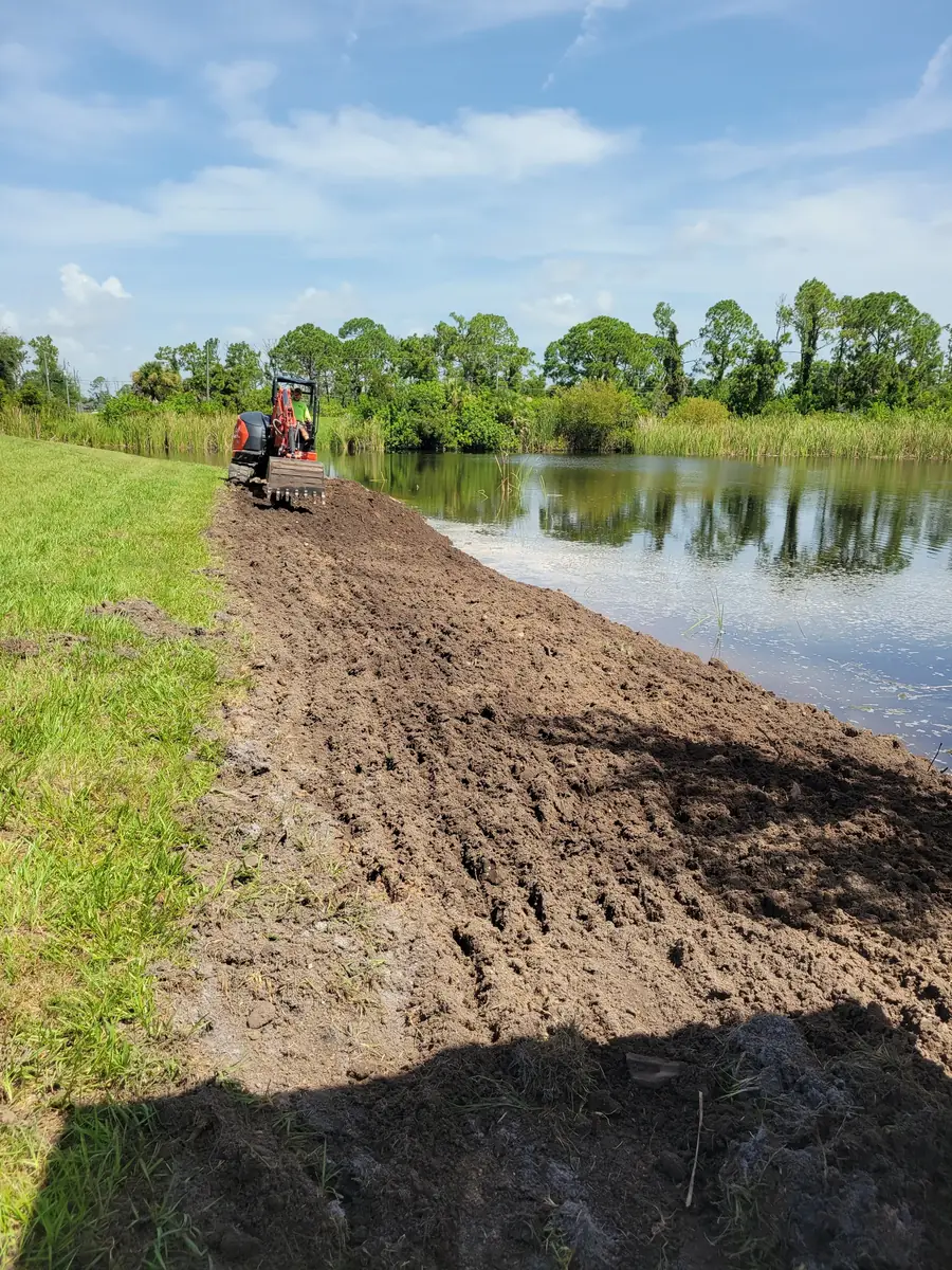 Mini excavator spreading new soil along a pond bank next to mowed turf