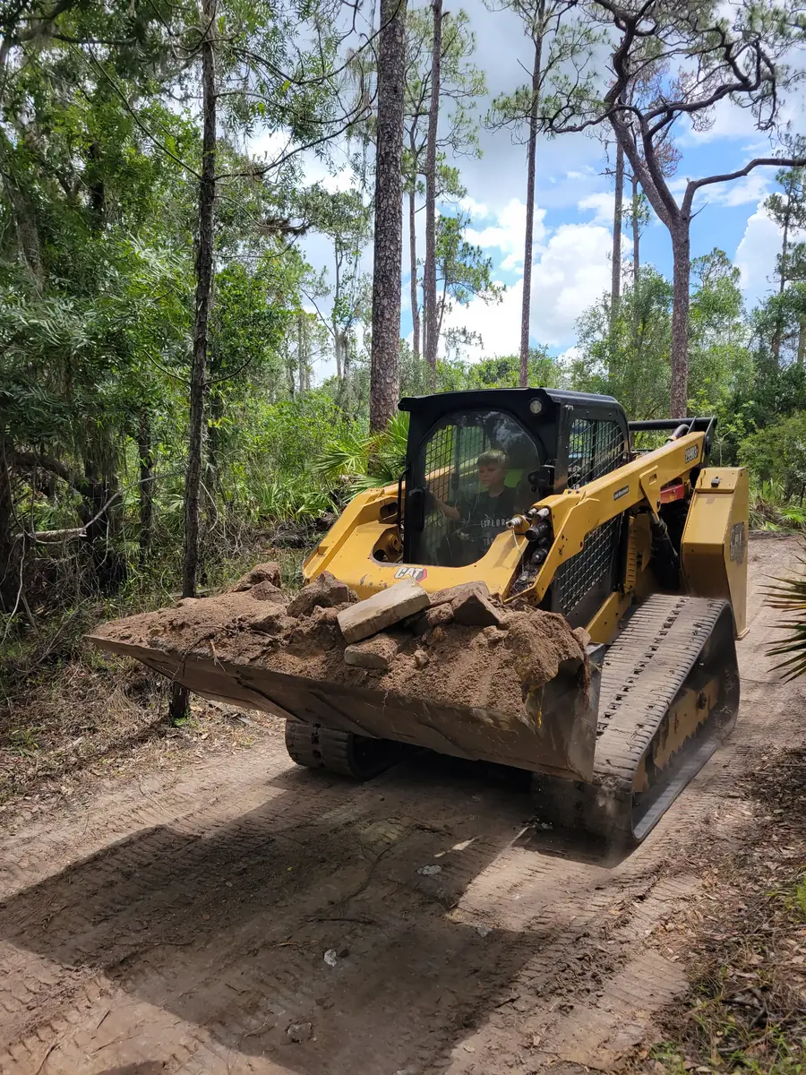 Tracked skid steer carrying a full bucket of soil down a sand trail through pines and palmetto