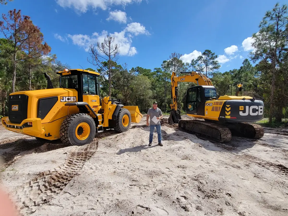 Operator standing between a wheel loader and a tracked excavator on a cleared sandy lot